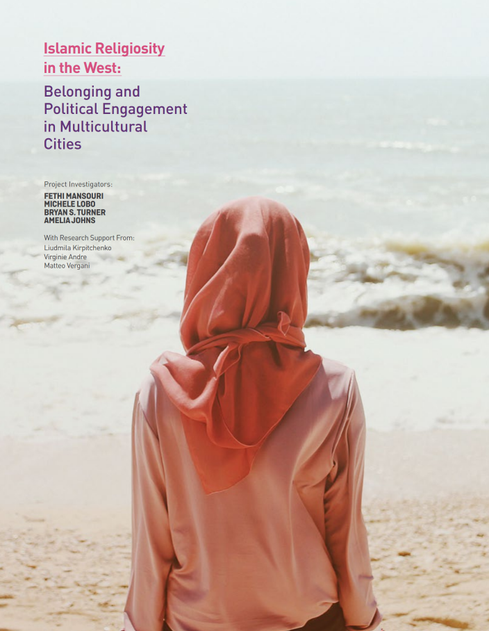 A woman wearing a peach coloured hijab and a lighter pink top, sitting on the sand at the beach in bright daylight. We see her from the back, facing the waves.
