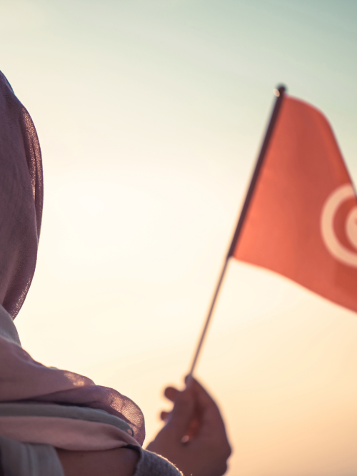 A woman wearing a peach coloured hijab and a lighter pink top, sitting on the sand at the beach in bright daylight. We see her from the back, facing the waves.