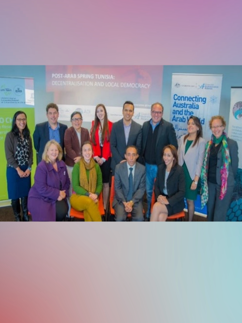 A woman wearing a peach coloured hijab and a lighter pink top, sitting on the sand at the beach in bright daylight. We see her from the back, facing the waves.