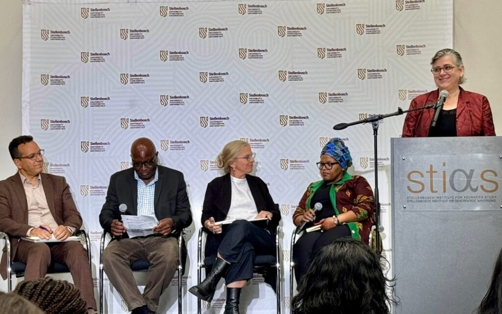 Four experts sitting on stage as part of a panel discussion at Stellenbosch University. The host stands at a podium next to the panel, introducing the event.