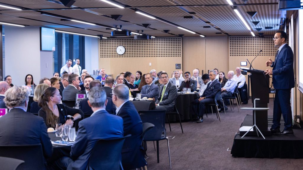 A man in a suit standing at a podium speaking to a room full of people seated at round dining tables